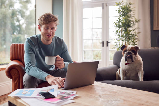 Man sitting in an armchair holding a mug, with a laptop on the table in front of him. On a nearby chair, a cute dog watches him