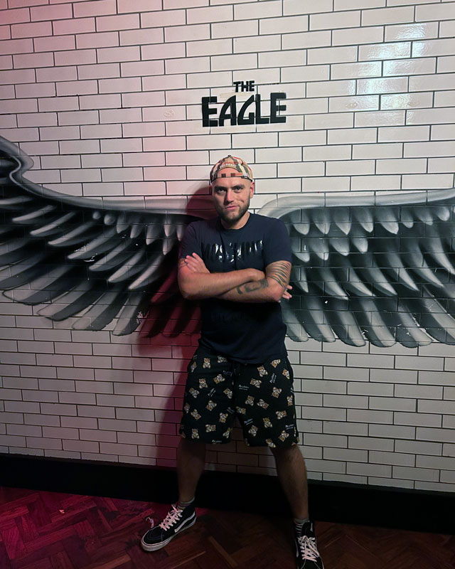 Person posing with arms crossed, standing against a wall with painted black wings, giving the illusion of having wings. Text above reads "The Eagle."