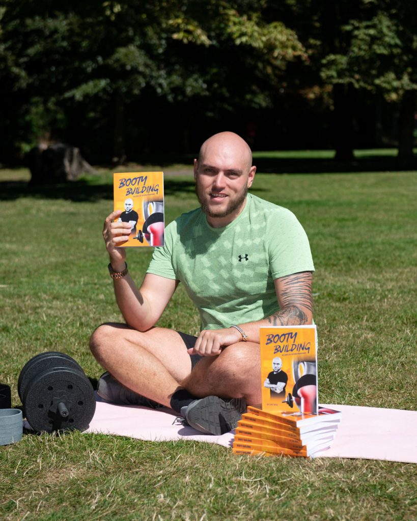 A man sits cross-legged on grass, with dumbbells and a pile of books nearby. He holds one copy of the book up to show the cover.