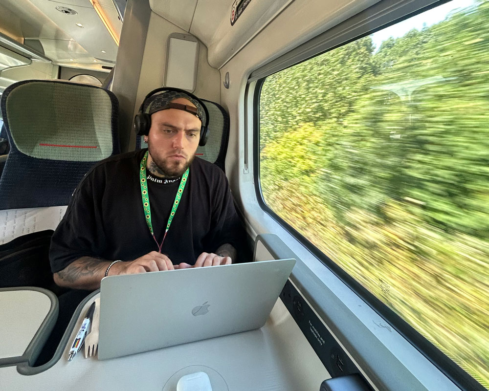A man wearing headphones and a sunflower lanyard works at a laptop on a train.