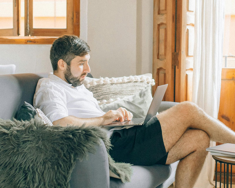 A man sits comfortably on a sofa, focused on a laptop. He wears a white shirt and black shorts, surrounded by soft pillows, exuding a relaxed, cozy ambiance.