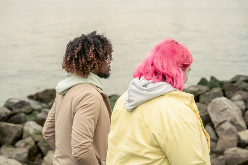 Two people stand on a rocky shoreline looking at the calm water. One has curly hair and a tan coat; the other, pink hair and a yellow jacket, suggesting contemplation.