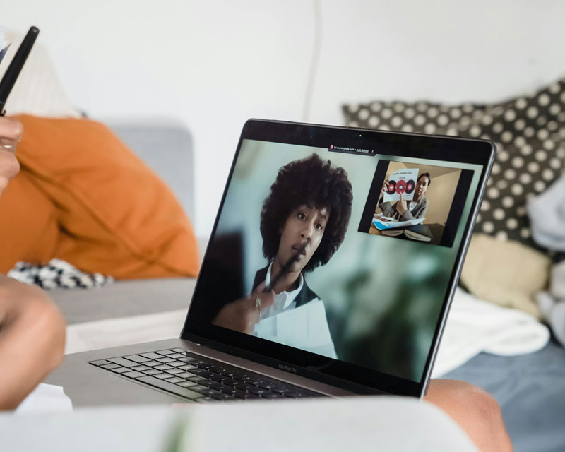 Laptop screen showing a video call with one person pensively holding a pen. Background shows a cozy room with orange and black cushions.
