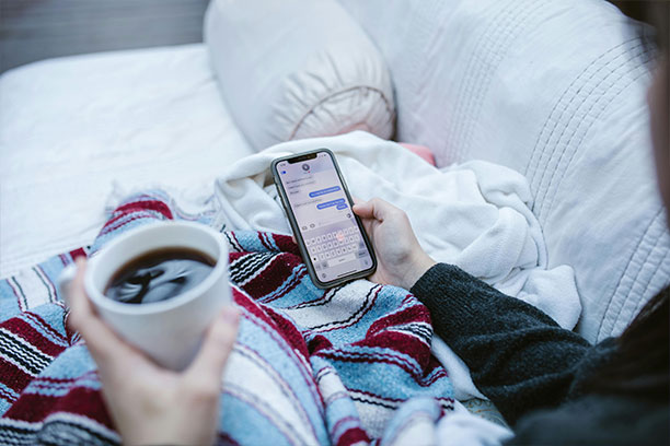 a person relaxes under a stripy blanket, holding a coffee in one hand, and with the other, using a messenger app on their phone.