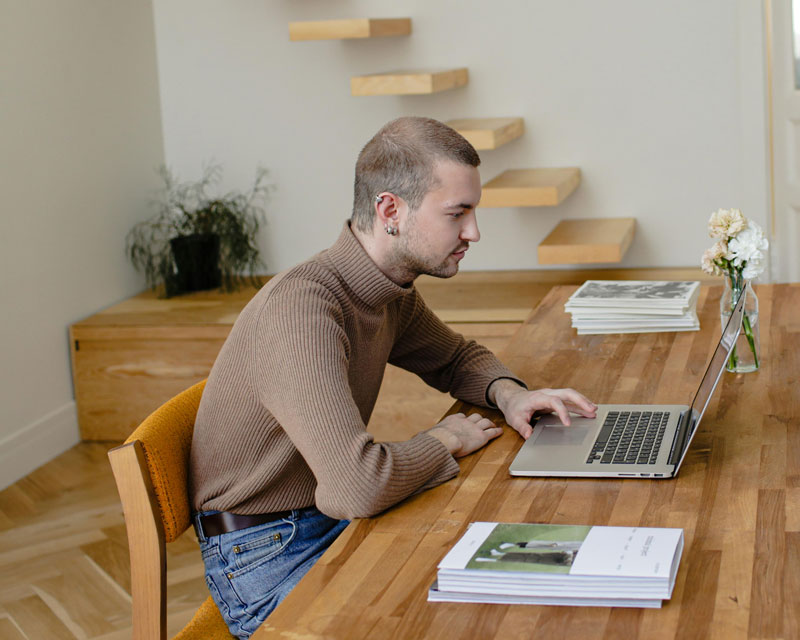 A person with short hair and a brown sweater works on a laptop at a wooden table. Books, a small vase with white flowers, and a minimalist wall shelf are visible.