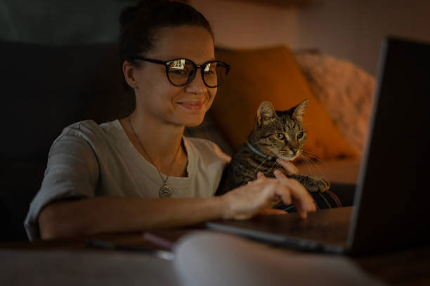 A woman with curly hair and glasses works at her laptop, while a cat cuddles up to her.