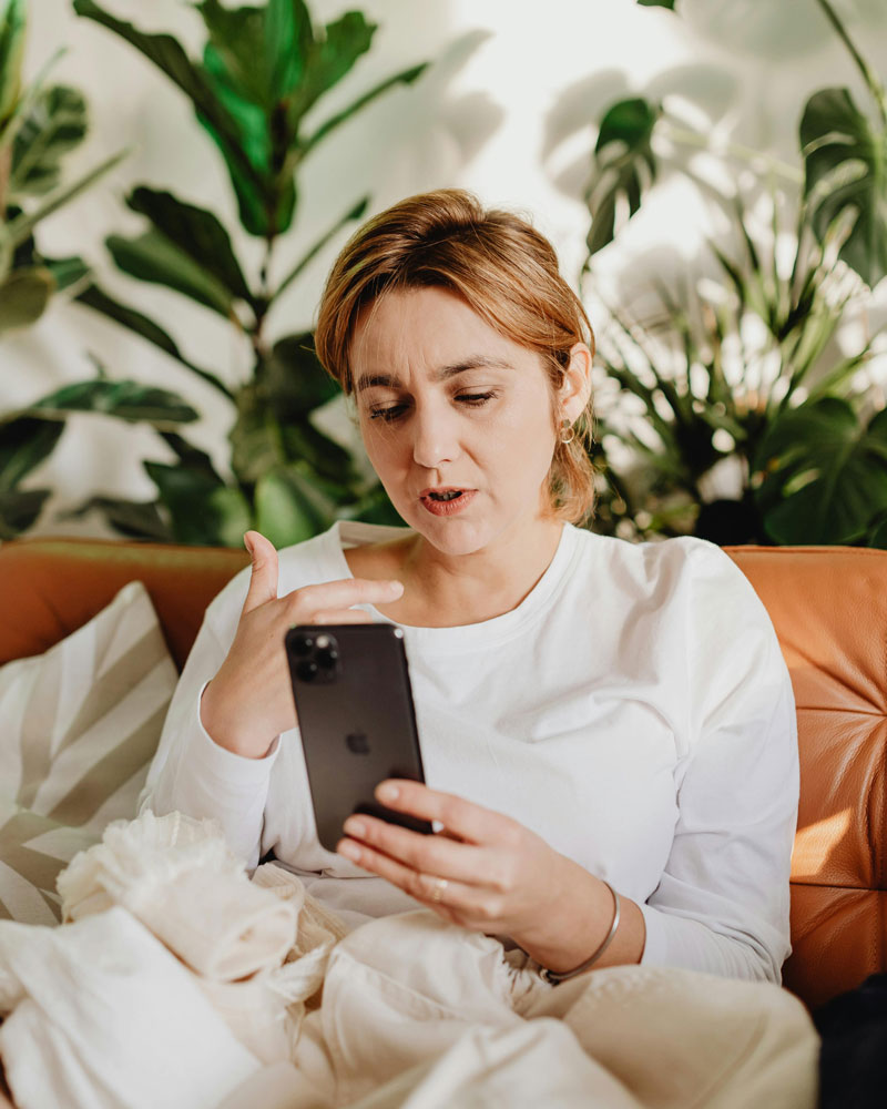 A woman in a white shirt sits on a couch, looking at a smartphone. She's surrounded by lush green plants, conveying a calm and focused mood.
