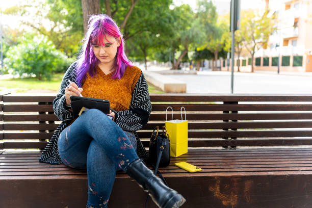 A woman with bright pink hair sits on a park bench while working on a tablet.