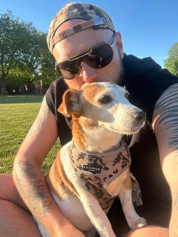 A man in a cap and sunglasses cuddles a dog wearing a camouflage harness in a sunlit park. He has tattoos, and they both look content and relaxed.