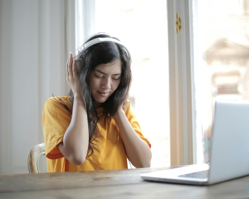 Woman in a yellow shirt enjoying music with white headphones, eyes closed, seated at a wooden table with a silver laptop, near a bright window.