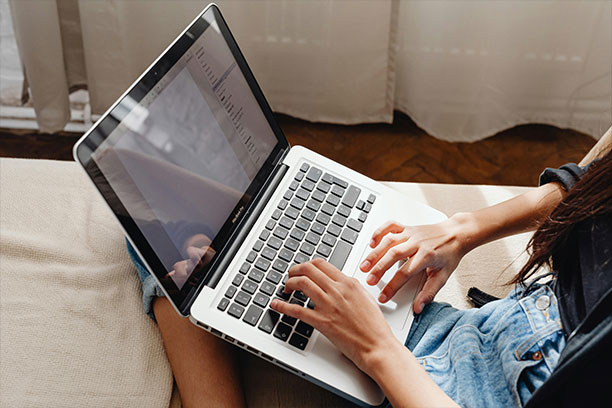 A person types on a laptop while sitting on a couch, conveying a relaxed and focused atmosphere. Sunlight filters through sheer curtains in the background.