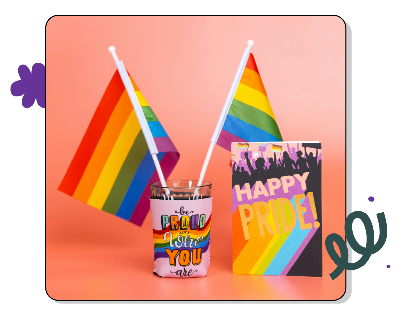 Two small rainbow flags, a colourful card saying "Happy Pride," and a glass with "Be Proud of Who You Are" text, set against a pink background.